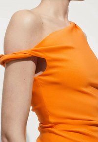 Person, die ein leuchtend orangefarbenes Top mit einem asymmetrischen Trägerdesign am Oberarm trägt, sichtbar vom Hals bis zur Taille.