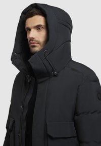 Dikke zwarte parka met een grote capuchon, ritssluiting aan de voorkant, drukknoopsluitingen en twee klepzakken. Gladde textuur met gestikte details en verstelbare bandjes.