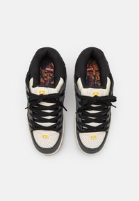Black and cream low-top sneakers with a textured finish, featuring a reinforced toe cap, black laces, and a patterned insole. Yellow logo accent.