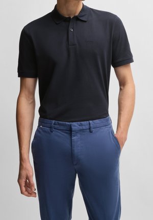 Homme portant un polo noir à manches courtes avec le logo "BOSS" et un pantalon bleu ajusté, les mains dans les poches.