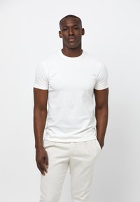 T-shirt en coton blanc avec un col rond, des manches courtes et une coupe ajustée, associé à un pantalon beige clair. Texture lisse, détails minimaux.