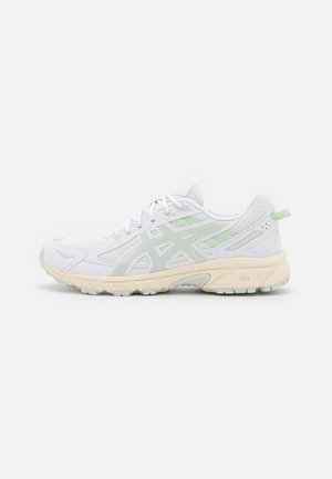 Weißer und hellgrüner Laufschuh mit Mesh-Obermaterial, gepolsterter beiger Sohle und Schnürung, von der Seite auf weißem Hintergrund gezeigt.