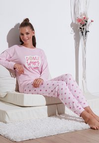 Rózsaszín pizsamakészlet, amely hosszú ujjú pólóból áll, rajta a "DONUT WORRY BE HAPPY" felirattal, és rózsaszín pöttyös nadrágból, egy modellel, aki párnán ül.