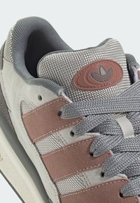 Adidas sneaker featuring a grey fabric upper, light grey suede accents, three pink stripes, and a textured grey tongue with logo.