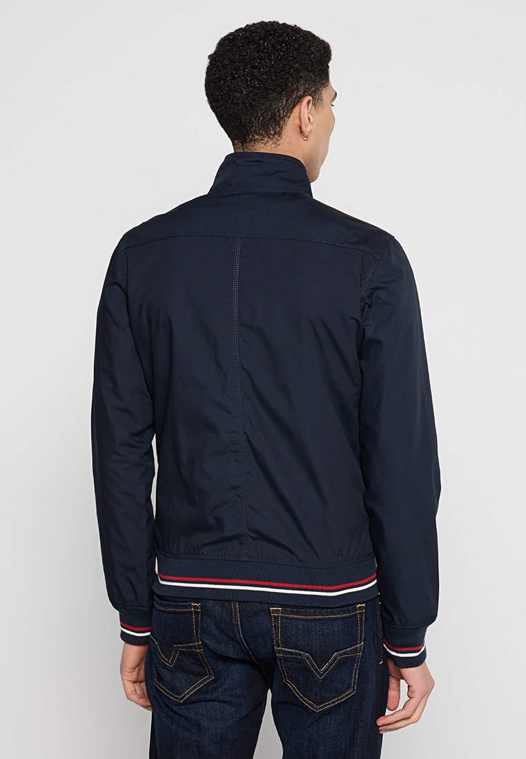 Veste zippée bleu marine avec un col montant, des poignets et un ourlet côtelés, ornée de rayures rouges et blanches, fabriquée en tissu léger.