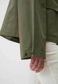 Giacca verde oliva in un tessuto leggero con una vestibilità ampia. I dettagli notevoli includono due bottoni decorativi sui polsini e maniche a taglio dritto.