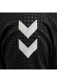 Svart sportströja med texturerad tyg och prickigt mönster. Notabelt vitt chevron-logga på ärmen, som kontrasterar mot det svarta.