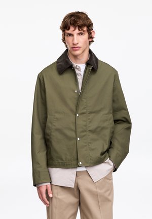 Jeune homme portant une veste vert olive avec un col foncé, une chemise beige en dessous et un pantalon marron clair, debout avec une main dans la poche.