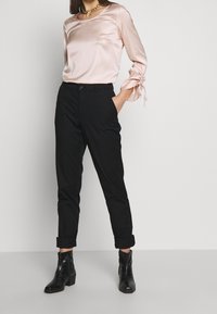 Ljust rosa satinblus med långa ärmar och knutna manschetter, kombinerad med svarta byxor och svarta ankelboots, vilket skapar en elegant look.