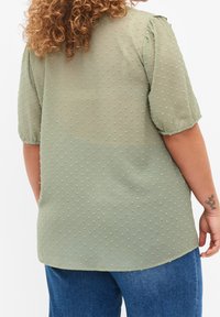 Blusa verde chiaro, tessuta, con maniche corte e un motivo a pois, abbinata a jeans blu. Il tessuto è morbido e leggermente trasparente.