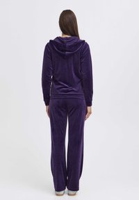Fialový velúrový loungewear set pozostávajúci z mikiny s kapucňou a širokých nohavíc. Mäkká textúra s hladkým povrchom a rebrované manžety na mikine.