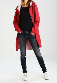 Rød vandtæt regnjakke med hætte, båret over en sort top og mørke jeans. Har trykknapper og et hvidt foret indre.