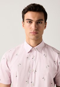 Chemise à manches courtes en coton rose clair avec un col boutonné, présentant un motif d'oiseaux et de fleurs aux couleurs atténuées.