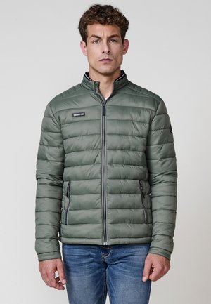 Chaqueta acolchada verde con patrón de acolchado horizontal, bolsillos con cremallera, cuello alto y acentos en negro contrastantes. Llevada sobre jeans de mezclilla.