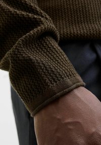 Hnědý pletený svetr se strukturou, s ribbovaným manžetovým lemem. Detailní snímek odhaluje pletený vzor a hladkou texturu pleti pod ním.