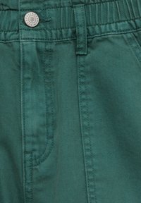 Primer plano de pantalones de mezclilla en color teal mostrando la cintura con elástico, la trabilla para el cinturón, el bolsillo delantero y un botón metálico etiquetado "Pull&Bear Denim".
