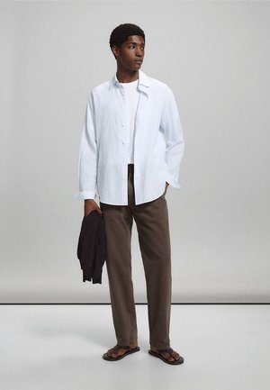 Jeune homme portant une chemise blanche texturée boutonnée sur un T-shirt blanc, pantalon marron, tongs noires, tenant une veste noire, debout sur un fond gris.