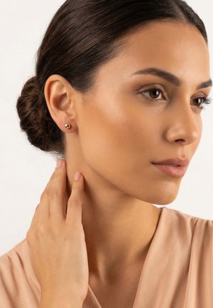 Kvinde med brunt hår i en knold iført små runde gyldne ørestikker, der rører ved halsen, iført en beige top mod en ensfarvet lys baggrund.