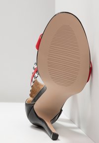 Sandale à talons hauts noire avec des brides rouges et à carreaux noir et blanc, vue de la semelle avec un motif antidérapant texturé.