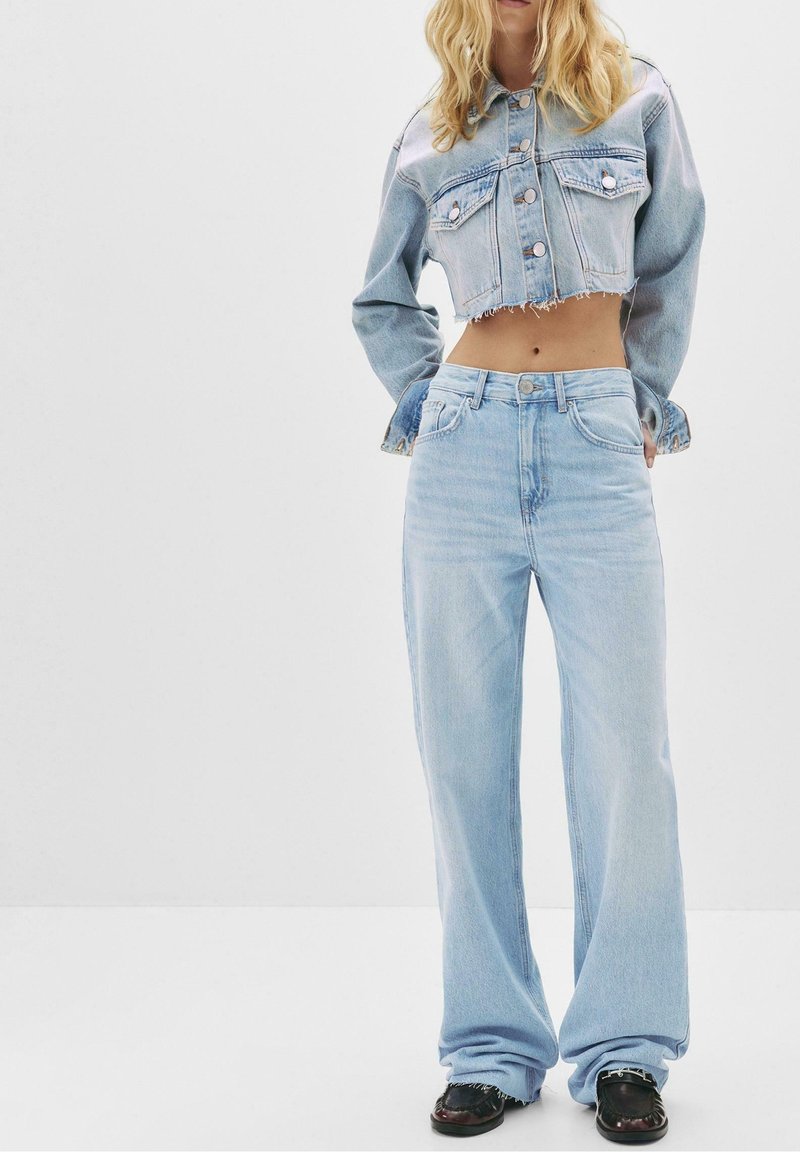 Femme portant une veste en jean courte délavée claire et un jean ample taille haute délavé clair, debout les mains dans les poches.
