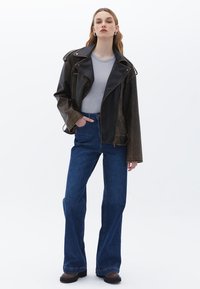 Hnědá a černá kožená motorkářská bunda přes šedé tričko, doplněná modrými rozšířenými džínami a černými masivními botami. Jednoduché bílé pozadí.
