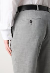 Pantaloni grigi su misura con una texture sottile, dotati di una tasca posteriore con un bottone nero e una cintura nera, indossati con una camicia bianca a maniche lunghe.