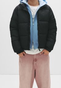 Sort dunjakke med blank finish, lynlåslukning og elastiske manchetter båret over en lyseblå hættetrøje, kombineret med lyserøde denim bukser.