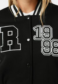 Černá varsity bunda se bílými pruhy na límci, cvokovými knoflíky a vyšívanými nášivkami: "R" a "1988" v kontrastních detailech.