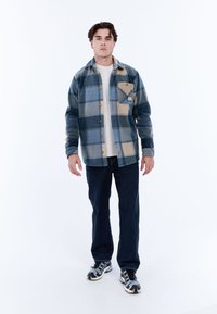 Jonge man die staat, gekleed in een blauw en beige geruit overhemd, een wit T-shirt, donkere jeans en zwart-witte sneakers op een witte achtergrond.
