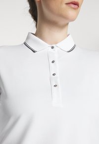 Biała koszulka polo z klasycznym kołnierzykiem, z czarnym paskiem wzdłuż krawędzi kołnierza i pięcioma metalowymi guzikami z przodu.