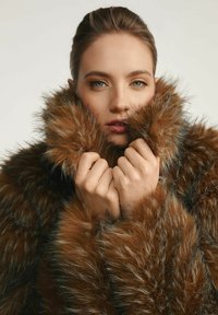 Brązowy płaszcz z sztucznego futra z grubym, teksturowanym kołnierzem. Modelka trzyma kołnierz, podkreślając miękkość płaszcza i bogate odcienie kolorów.