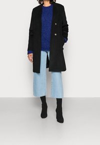 Černý kabát s propracovaným střihem a zlatými knoflíky, vrstvený přes modrý pletený svetr a světlé vysoko pasové džíny. Černé kotníkové boty.