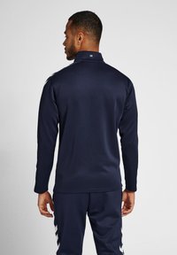 Maglietta sportiva a maniche lunghe color navy con colletto alto, texture liscia e dettaglio del logo bianco sul retro, abbinata a pantaloni dello stesso colore.
