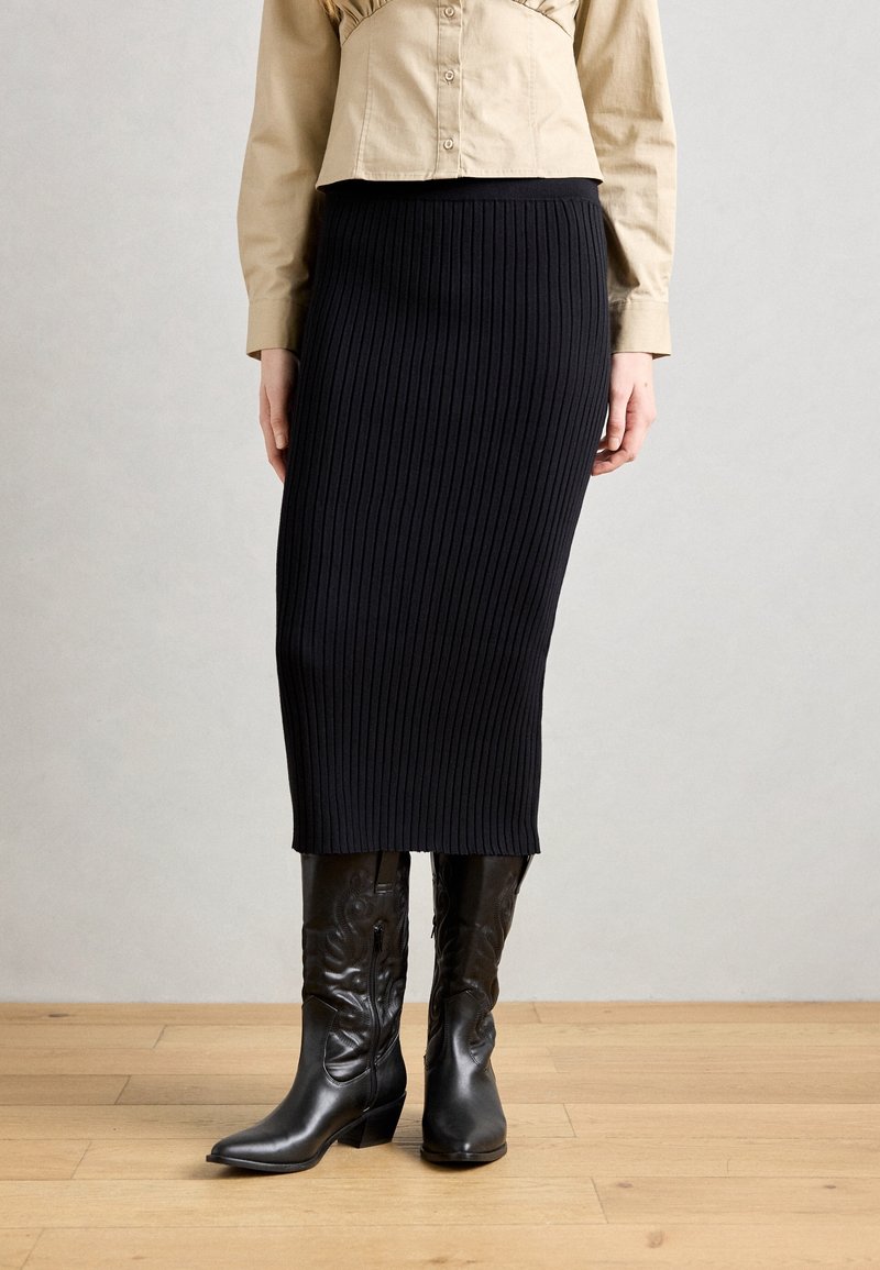 Femme portant une chemise beige boutonnée, une jupe midi côtelée noire et des bottes de cowboy en cuir noir, debout sur un sol en bois.