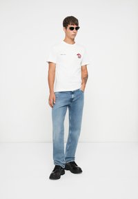 White cotton t-shirt with logo print, paired with light blue jeans, and black chunky shoes. Model wears black sunglasses and has short hair.