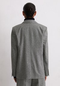 Blazer en laine grise à la coupe ajustée, col à revers cranté et poignets boutonnés. Tissu texturé avec un motif subtil. Vue arrière montrant une couture centrale.