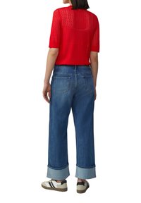 Rotes gestricktes Oberteil mit kurzen Ärmeln, kombiniert mit weiten blauen Jeans mit umgekrempelten Säumen. Sneakers mit einer Kombination aus hellen und dunklen Farben.