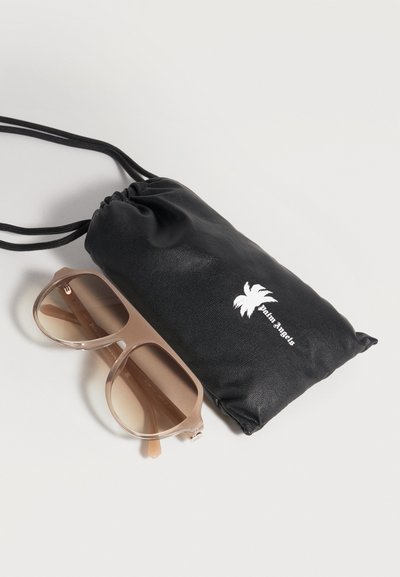 Lunettes de soleil carrées beige oversize avec verres dégradés à côté d'une pochette noire à cordon avec un logo palmier blanc.