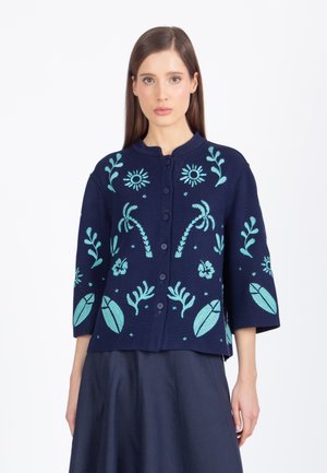 Kobieta o prostych brązowych włosach, ubrana w granatowy rozpinany kardigan z jasnoniebieską kwiatową i liściastą haftowaną ozdobą, w połączeniu z ciemną spódnicą.