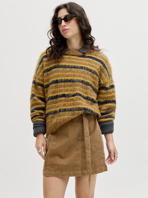 Kvinne med mørkt, bølget hår som har på seg sorte solbriller, en myk, stripete genser i sennepsgul og marineblå over en grå skjorte, og et sandfarget kordfløyelskjørt med knytebelte.