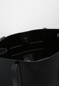 Svart läder tote-väska med tryckknappsstängning, inre fickor och tygetikett som visar en liten färgglad logotyp.