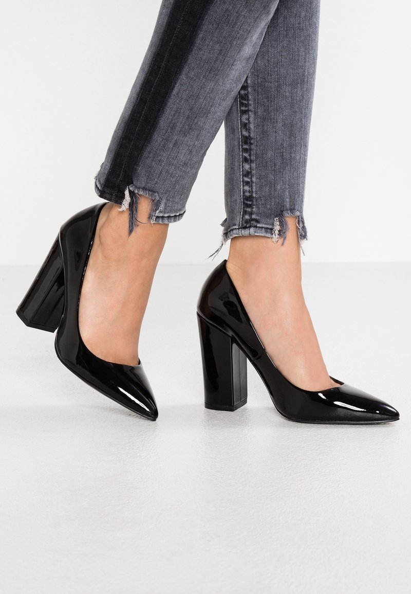 Pieds portant des talons blocs en cuir verni noir à bouts pointus, associés à un jean skinny gris à ourlet effiloché.