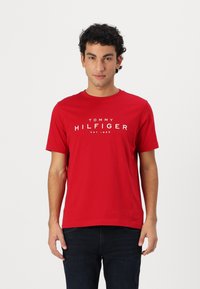 Raudona medvilninė marškinė su trumpomis rankovėmis, apvaliu iškirpimu ir baltu "TOMMY HILFIGER" logotipu krūtinėje. Lygus tekstūra.