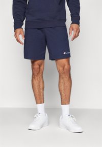 Pantalones cortos de algodón azul marino con una cinturilla elástica, que presentan un pequeño logo blanco de Champion en el dobladillo, combinados con zapatillas deportivas blancas y calcetas.