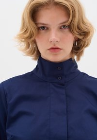 Giovane con capelli biondi ondulati e corti, che indossa orecchini a cerchio argentati e una camicia blu navy con colletto alto, rivolto verso l'osservatore.