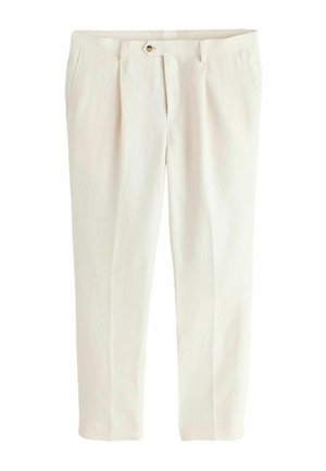 Mand iført cremefarvet jakkesæt, mørk skjorte, solbriller og sorte sandaler står indendørs nær en beige sofa og et glasvindue, der reflekterer træer.