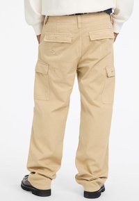Beżowe spodnie cargo o luźnym kroju, z wieloma kieszeniami, delikatnymi plamami farby i mankietami na dole. Tkany bawełniany materiał.