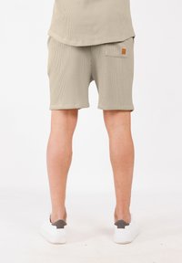 Personne portant un short texturé beige et un haut assorti, des baskets blanches, se tenant contre un fond blanc uni, vue de dos.