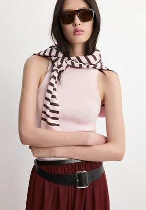 Donna con capelli lunghi e scuri che indossa grandi occhiali da sole marroni, top rosa a coste senza maniche, sciarpa a righe legata sulle spalle, gonna bordeaux e cintura nera.