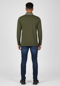 Man som står med ryggen vänd, iklädd en olivgrön långärmad tröja, blå jeans och svarta sneakers mot en enfärgad bakgrund.